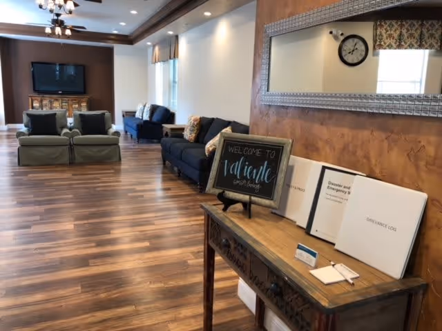 Communal living room with sofas, a wall-mounted TV, wood floors, and a console table displaying a "Welcome to Valiente Senior Living" sign.