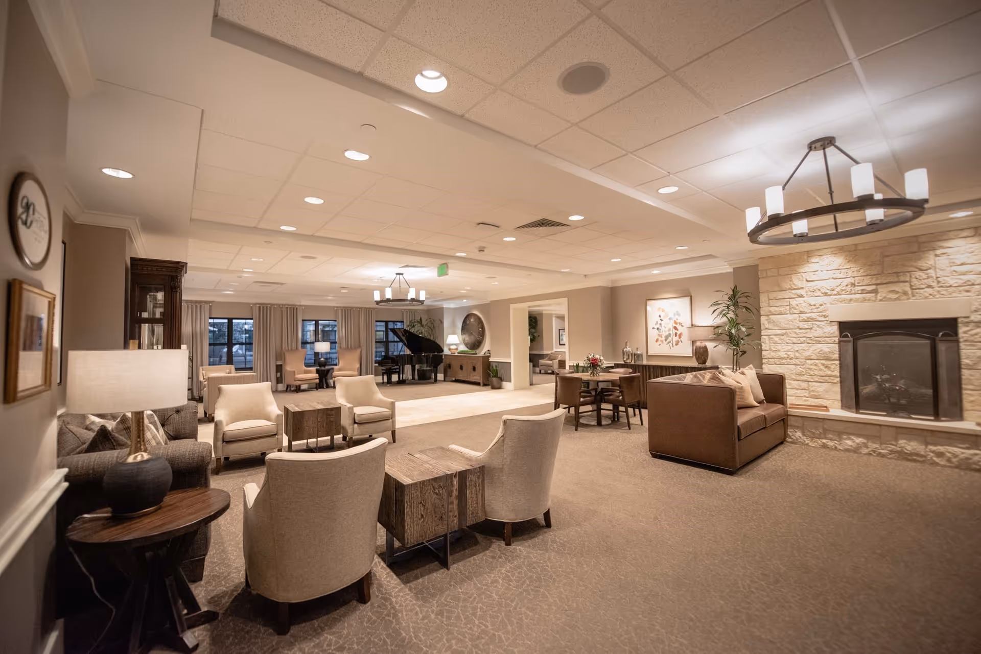 A spacious and well-lit senior living facility common area with comfortable seating including armchairs and sofas arranged around small tables. The room features a stone fireplace on the right wall, a grand piano in the background near large windows with curtains, and warm lighting from ceiling fixtures and lamps. The decor includes framed artwork, plants, and neutral tones creating a cozy and inviting atmosphere.