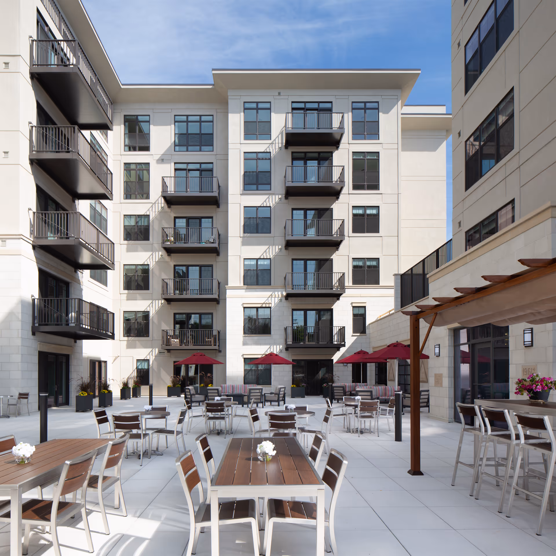 Outdoor patio area of St. Rita Square with multiple tables and chairs arranged for seating. The space includes red umbrellas, a pergola with bar stools, and is surrounded by a multi-story building with balconies and large windows under a clear sky.