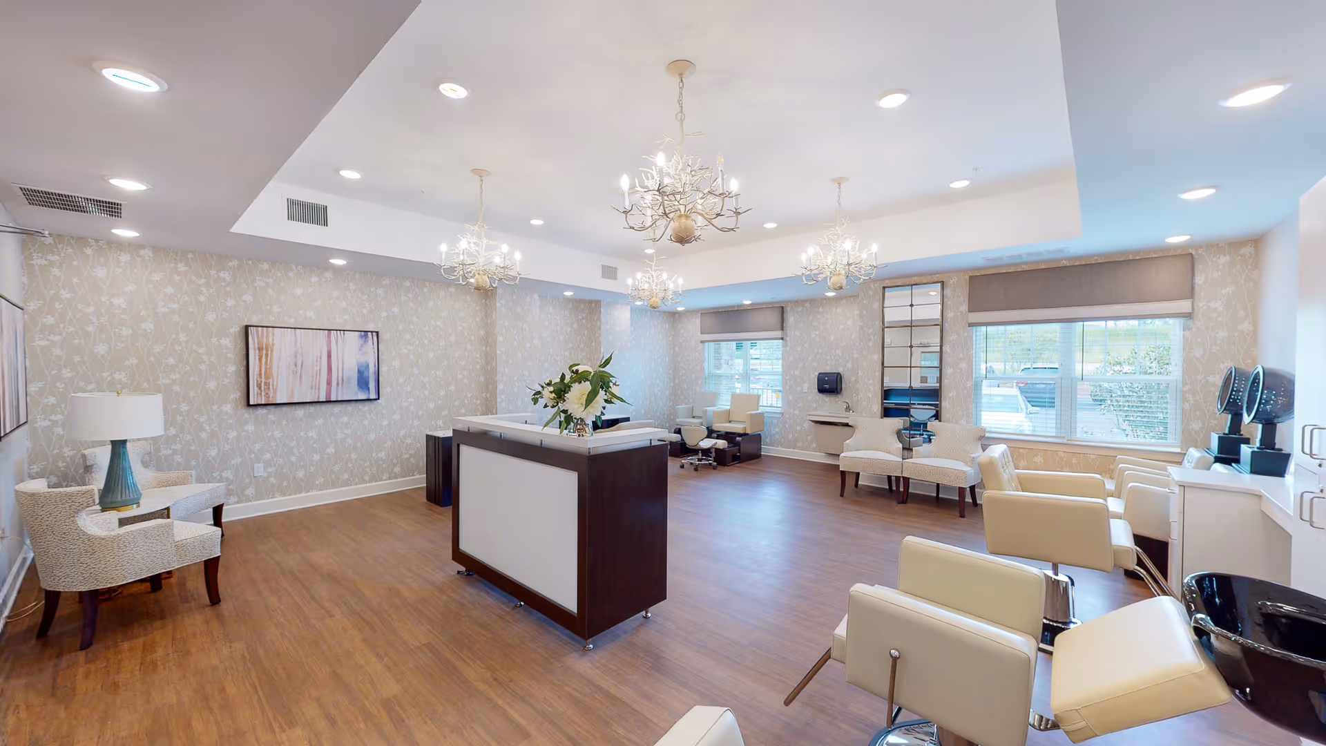 A spacious and well-lit salon area with multiple white salon chairs, a central reception desk with a flower arrangement, decorative chandeliers, patterned wallpaper, and large windows allowing natural light to enter.