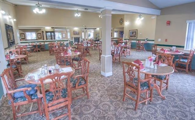 A spacious dining room with multiple round wooden tables and chairs arranged neatly. Each table is set with napkins, glasses, and small flower vases. The room has carpeted flooring with a patterned design, beige walls, and several light fixtures on the ceiling. Large mirrors and framed pictures decorate the walls, and windows allow natural light to enter.