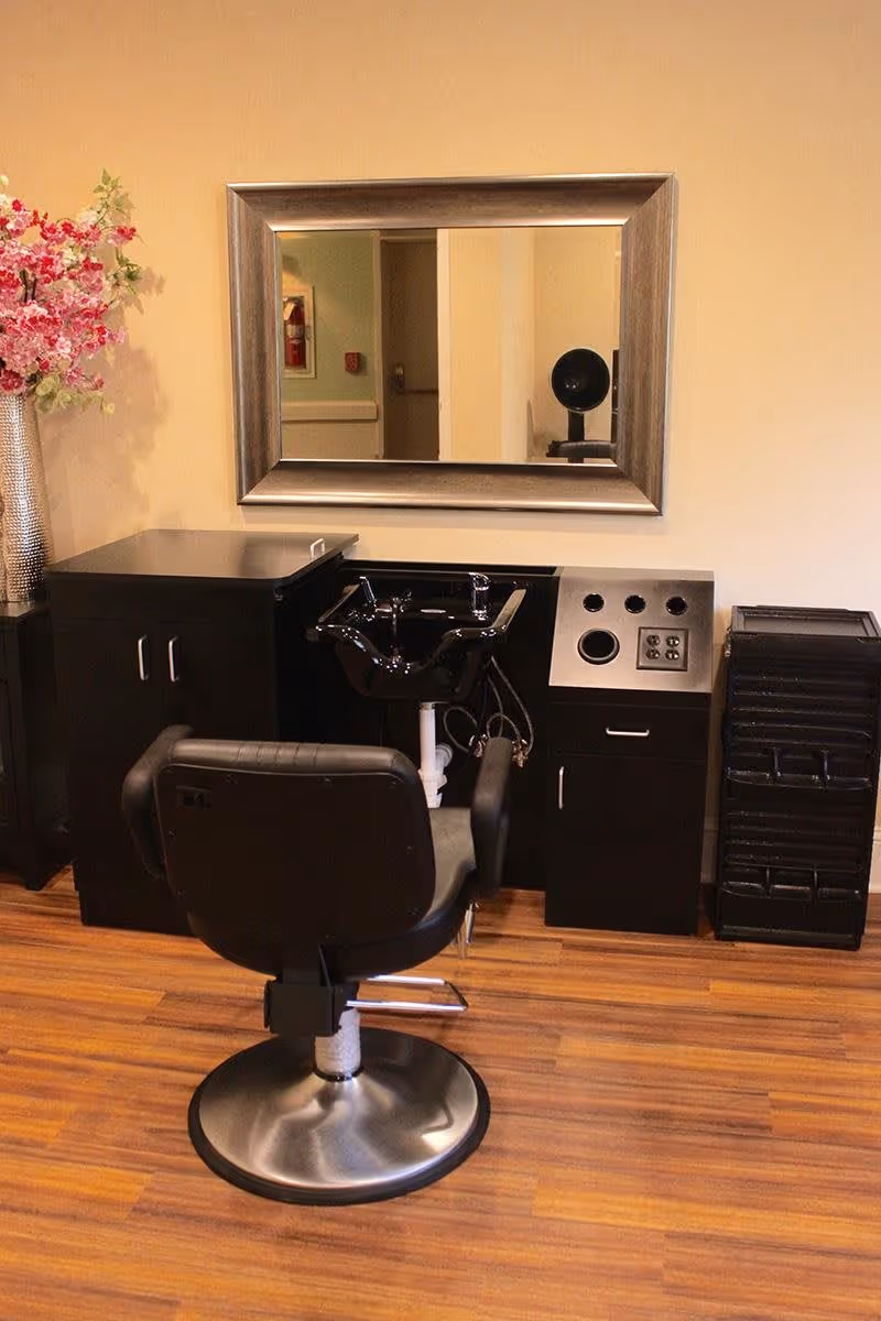 A salon station in a senior living facility with a black salon chair in front of a black sink and cabinetry. A large rectangular mirror with a silver frame is mounted on the wall above the sink. To the left, there is a tall vase with pink flowers. The floor is wooden.