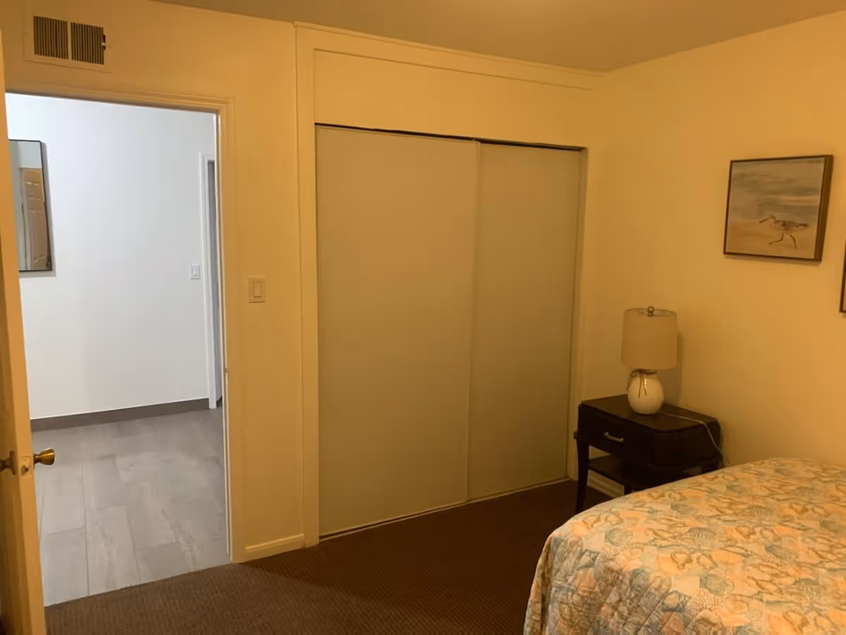 Small bedroom with a bed, nightstand and lamp, sliding closet doors, and an open doorway to a hallway.