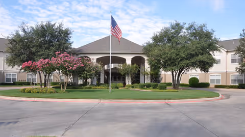 Front exterior view of Brookdale Eden Estates building with a circular driveway, manicured lawn, trees, and an American flag on a flagpole in the center.