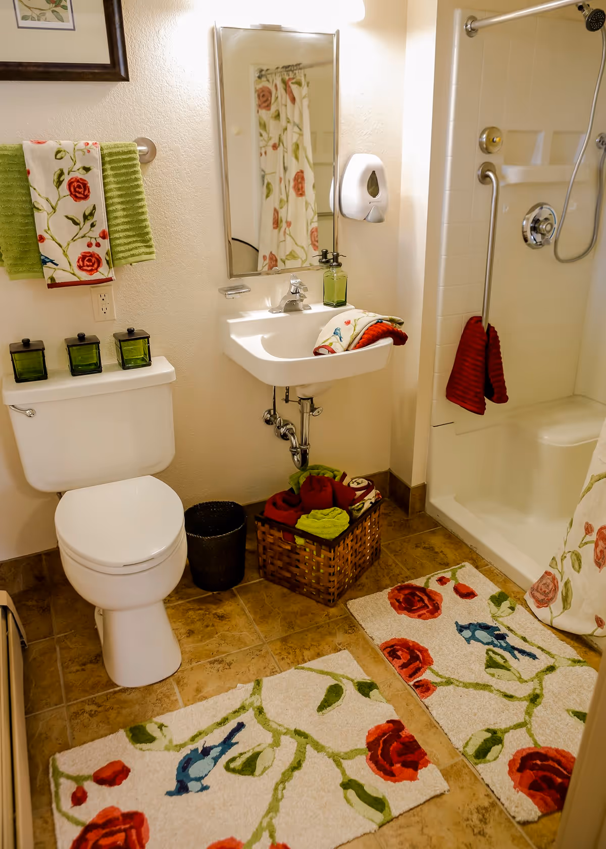 A clean and well-lit bathroom featuring a white toilet, a wall-mounted sink with a mirror above it, and a walk-in shower with a grab bar. The bathroom is decorated with floral-patterned towels, matching bath mats with red roses and green leaves, and a wicker basket filled with folded towels. The floor is tiled in a warm brown tone.