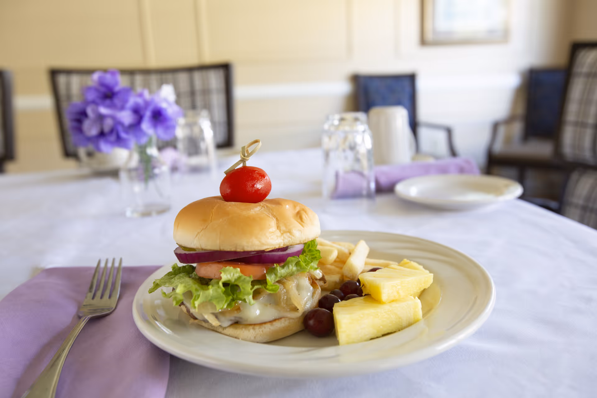 A plate with a cheeseburger topped with lettuce, tomato, and onion, accompanied by French fries, pineapple slices, and grapes, set on a white tablecloth with a purple napkin and fork. In the background, there are chairs and a small vase with purple flowers on the table.