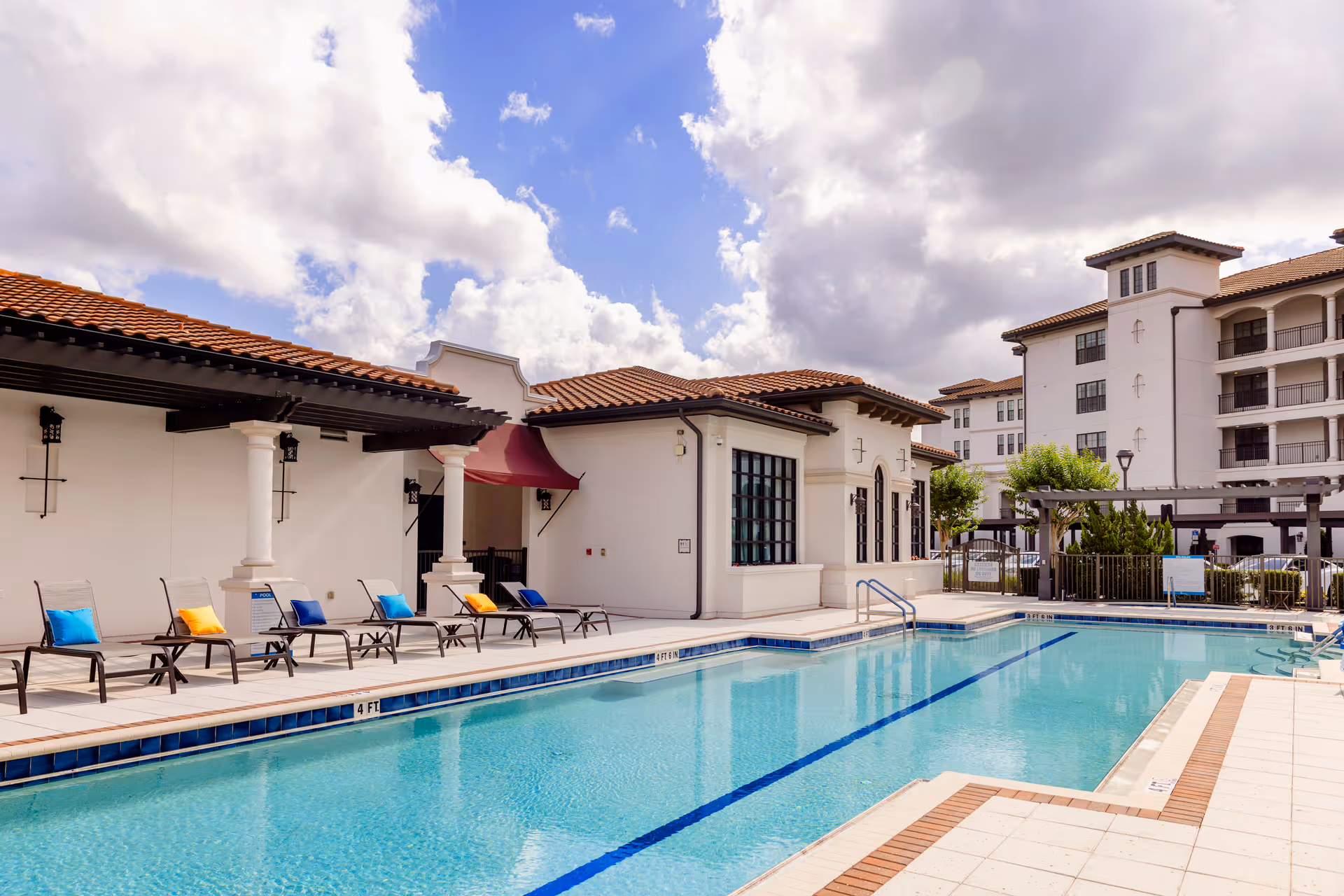 Outdoor swimming pool area at Westminster Baldwin Park with lounge chairs featuring colorful cushions along the poolside, adjacent to a building with white walls and red-tiled roof under a partly cloudy sky.