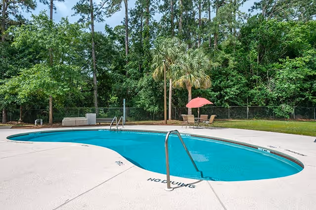Outdoor swimming pool with clear blue water surrounded by a concrete deck. There are metal handrails for entering the pool, a few lounge chairs, a table with an umbrella, and lush green trees and bushes in the background.