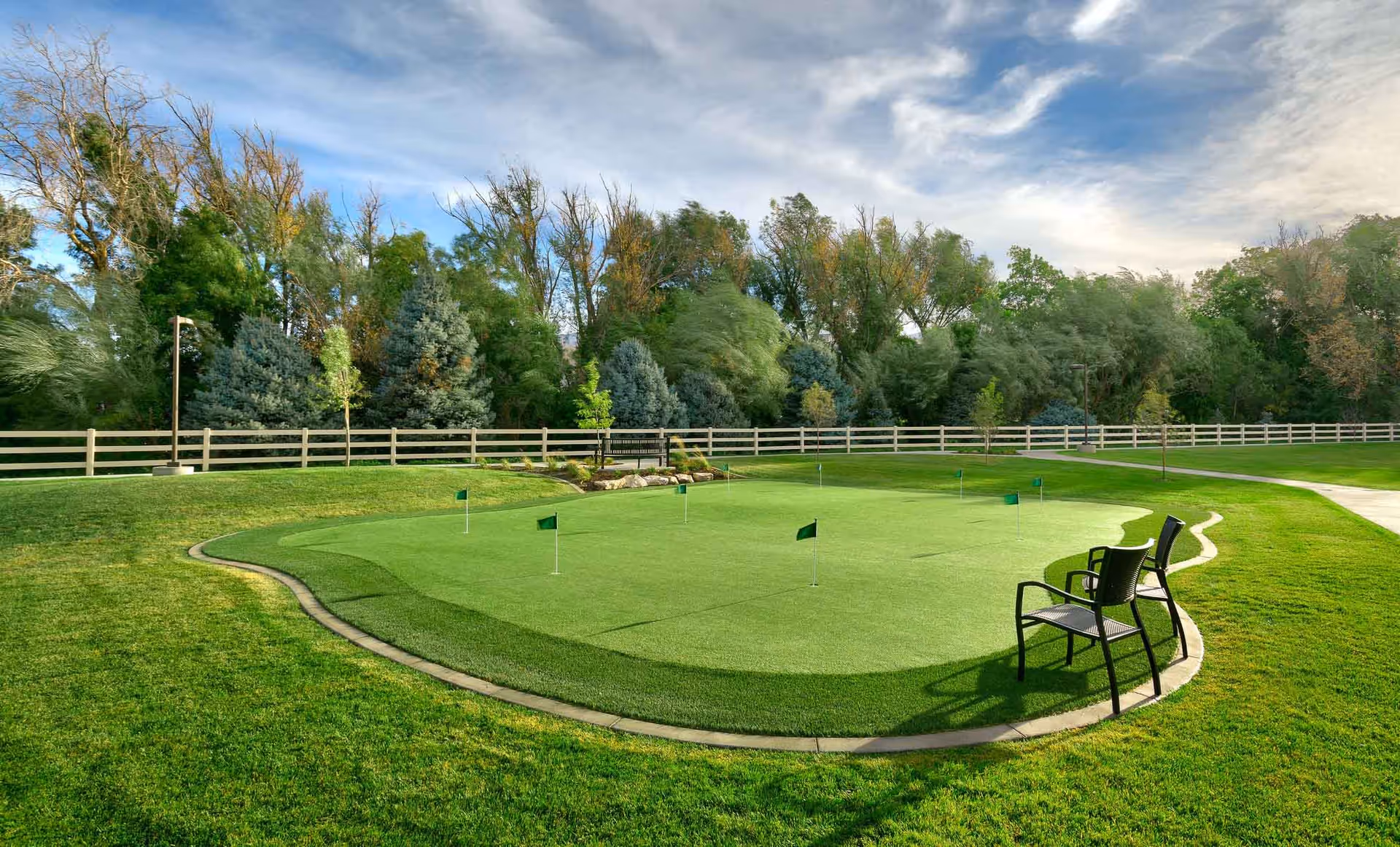 A landscaped outdoor putting green with several small flags, a pair of chairs, surrounding lawn, fence, and trees under a partly cloudy sky.