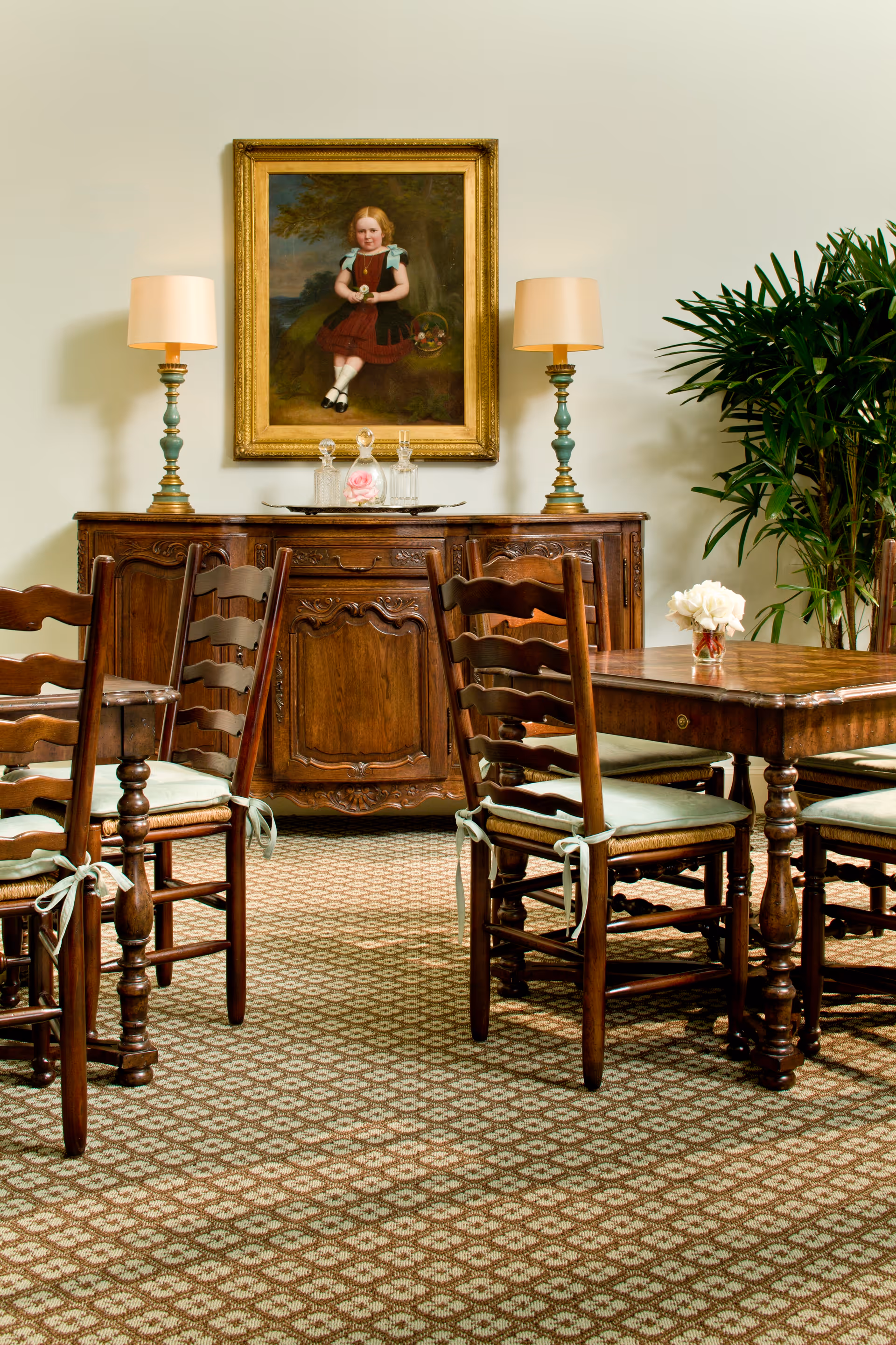 A traditional dining room with a wooden dining table and six wooden chairs with light green cushions. Behind the table is an ornate wooden sideboard with two matching lamps and glass decanters on top. Above the sideboard hangs a framed portrait of a young girl in a red dress holding a basket. To the right of the sideboard is a large green potted plant. The floor is covered with a patterned carpet.