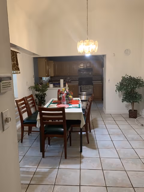 A dining area with a rectangular table covered with a white tablecloth and green placemats, surrounded by six wooden chairs with green cushions. A floral centerpiece and some items are on the table. The dining area is adjacent to a kitchen with wooden cabinets, a stove, and a tiled backsplash. A hanging light fixture is above the table, and there is a large potted plant on the right side near a white wall with a clock.