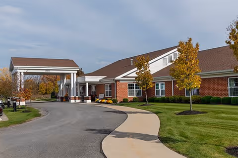 Exterior view of a single-story brick building with a covered entrance driveway, surrounded by a well-maintained lawn and young trees with autumn foliage under a cloudy sky.