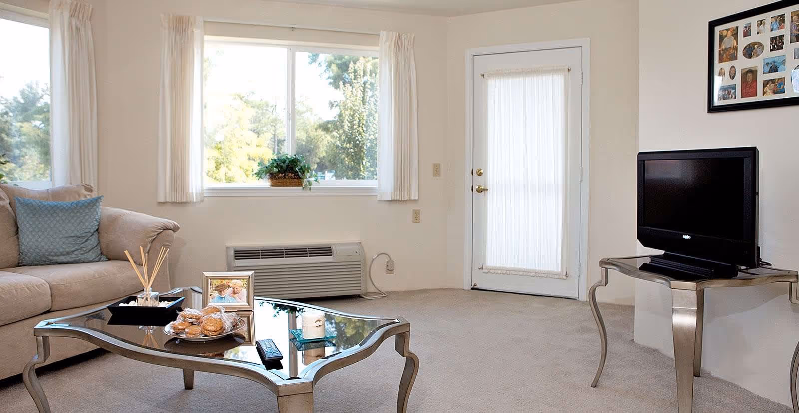 A bright living room with a beige sofa adorned with a blue cushion, a glass coffee table holding a plate of cookies, a framed photo, a candle, and a remote control. There is a large window with white curtains letting in natural light, a small plant on the windowsill, a wall-mounted air conditioning unit below the window, and a door with a sheer curtain. A flat-screen TV sits on a metallic side table, and a framed collage of photos hangs on the wall above it.