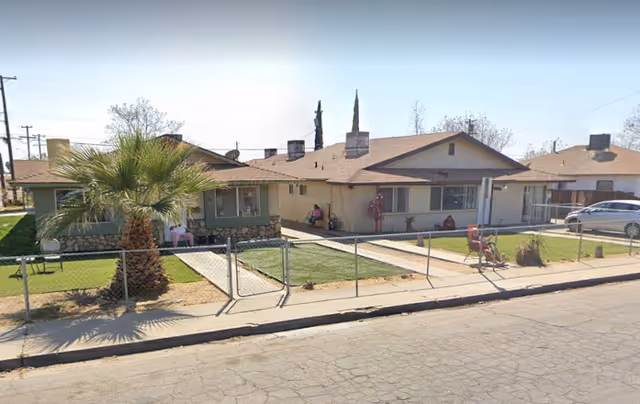 Single-story care home with a fenced front lawn, palm tree, and residents seated outside.