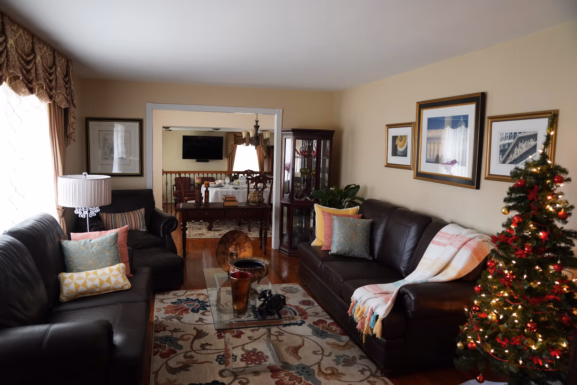 A cozy living room with two dark leather sofas adorned with colorful pillows and a throw blanket. A glass coffee table sits on a patterned rug in the center. There is a decorated Christmas tree with lights and ornaments on the right side. The room has beige walls with framed pictures and a window with draped curtains on the left. In the background, a dining area with a table set for a meal and a wall-mounted TV is visible.