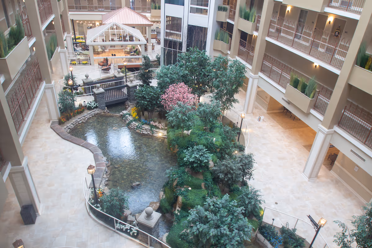 Indoor atrium area in a senior living facility featuring a small pond with clear water, surrounded by lush greenery and flowering plants. There is a small bridge over the pond and a white gazebo structure in the background. The area is enclosed by multiple floors with balconies and railings, and the floor is tiled with light-colored tiles.