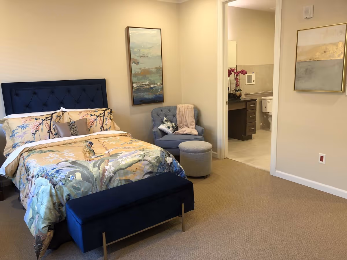 A bedroom with a bed featuring a dark blue tufted headboard and floral bedding. There is a matching dark blue bench at the foot of the bed. Next to the bed is a blue upholstered chair with a throw blanket and decorative pillow, accompanied by a round ottoman. The room has beige walls with two framed paintings and beige carpet. An open doorway reveals a bathroom with a vanity, mirror, and toilet.