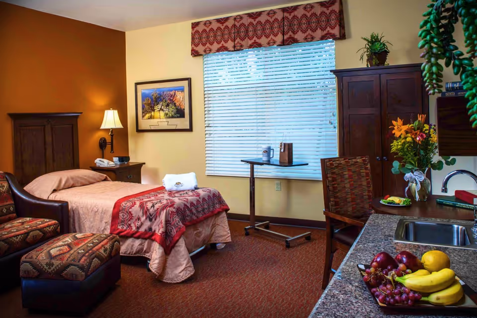 A cozy bedroom in a senior living facility featuring a single bed with a patterned red and beige bedspread, a wooden headboard, and a bedside table with a lamp and telephone. There is a comfortable armchair with a matching ottoman, a window with blinds and a decorative valance, a small adjustable table, a wooden wardrobe, and a countertop with a sink and a tray of fresh fruit including bananas, grapes, and apples. A vase with flowers and some books are also on the countertop.
