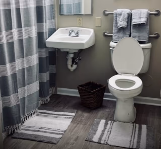 A bathroom with a white toilet with the lid open, a white wall-mounted sink with a faucet, a mirror above the sink, a striped shower curtain, two striped bath mats on the floor, a small dark woven basket, and two gray towels hanging on a grab bar behind the toilet.