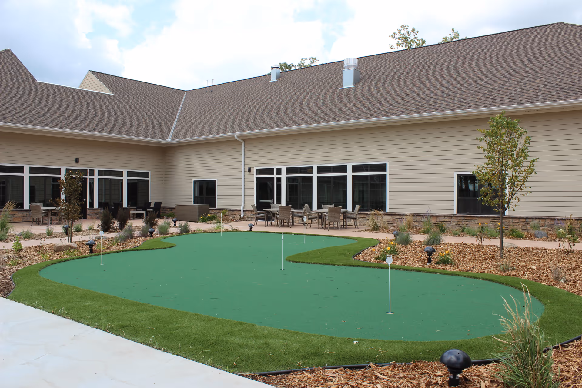 Outdoor courtyard area at Chateau Waters featuring a putting green with several golf holes, surrounded by landscaped mulch beds, small trees, and shrubs. There are multiple tables and chairs on the patio adjacent to the building with large windows and beige siding under a brown shingled roof.
