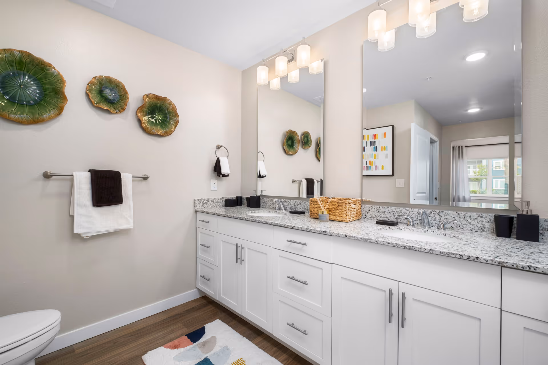 A modern bathroom with a white double-sink vanity topped with a speckled granite countertop. Above the sinks are two large mirrors with three-light fixtures. On the left wall, there are three decorative green and brown ceramic plates and a towel rack holding white and dark brown towels. The floor has wood-like flooring and a colorful patterned rug. A toilet is partially visible in the lower left corner.