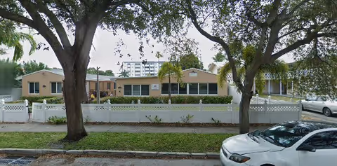 Exterior view of a single-story retirement home building with beige walls and a white fence in front. There are large trees and parked cars along the street in front of the building.
