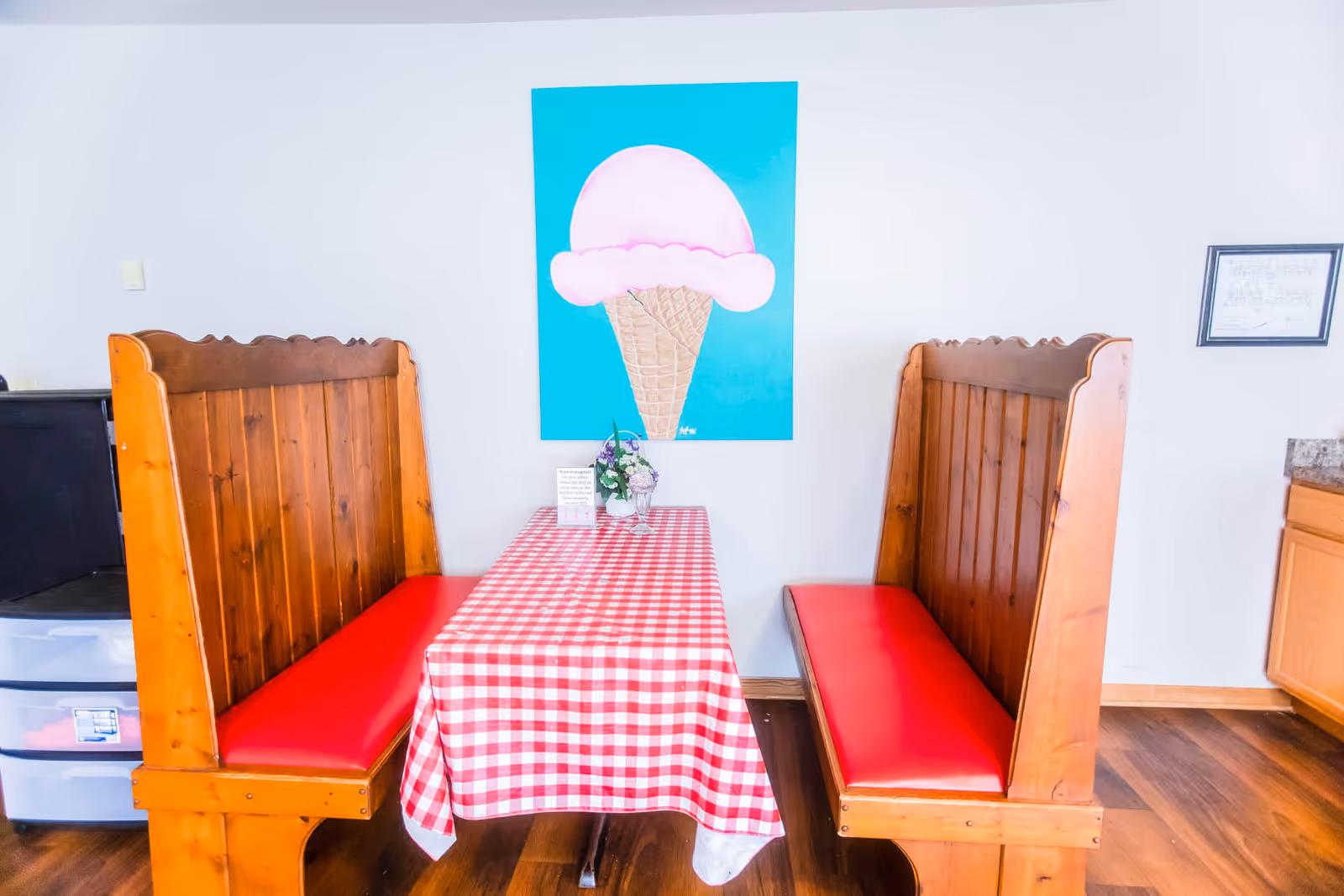 A dining area with two wooden booths featuring red cushioned seats facing each other across a table covered with a red and white checkered tablecloth. On the table, there is a small flower arrangement and a sign. A large painting of a pink ice cream cone on a blue background hangs on the wall behind the table. The floor is wooden, and part of a kitchen counter is visible on the right side.