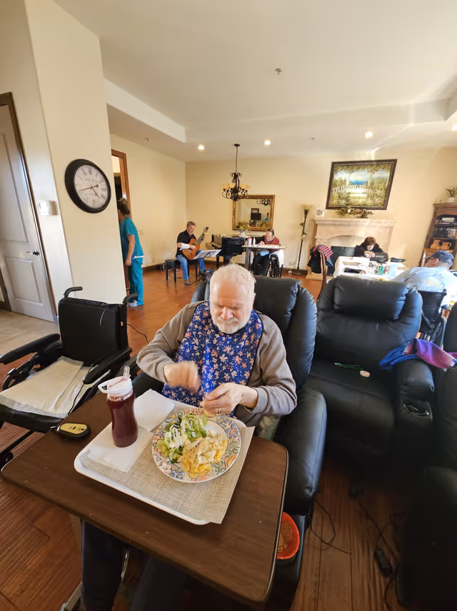 An elderly man wearing a floral bib sits in a recliner chair at a small table with a plate of food and a drink in front of him. Behind him, several other elderly individuals are seated at tables or in chairs in a spacious, well-lit room with wooden floors, a fireplace, and a large framed painting on the wall. A person in scrubs is walking near a doorway on the left side of the image.