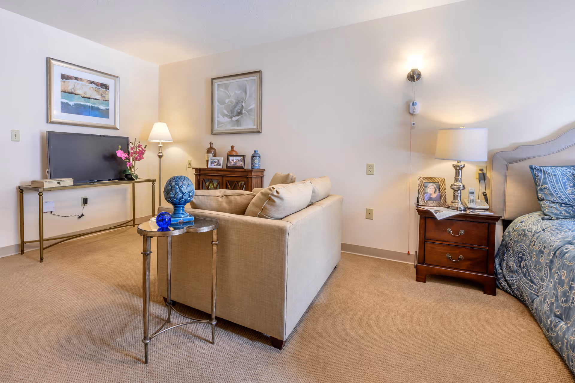 A cozy living space featuring a beige sofa facing a flat-screen TV on a slim metal stand, a small round side table with blue decorative items, a wooden nightstand with a lamp, framed photo, and a cordless phone, and a bed with blue patterned bedding partially visible on the right. The walls are adorned with framed artwork and the room is carpeted in a neutral tone.
