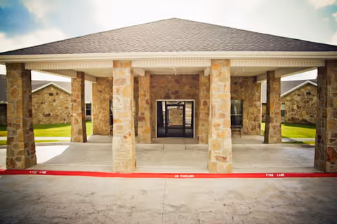 Front exterior view of a building with stone pillars supporting a covered entrance. The building has a stone facade and a dark shingled roof. There is a red painted curb in front marked with 'FIRE LANE NO PARKING'.