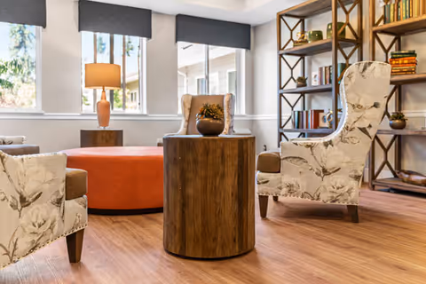 A cozy living room area with two floral upholstered armchairs facing a round wooden side table. In the background, there is a large orange ottoman, a wooden side table with a lamp, and a tall bookshelf filled with books and decorative items. Large windows with gray valances allow natural light to brighten the space.