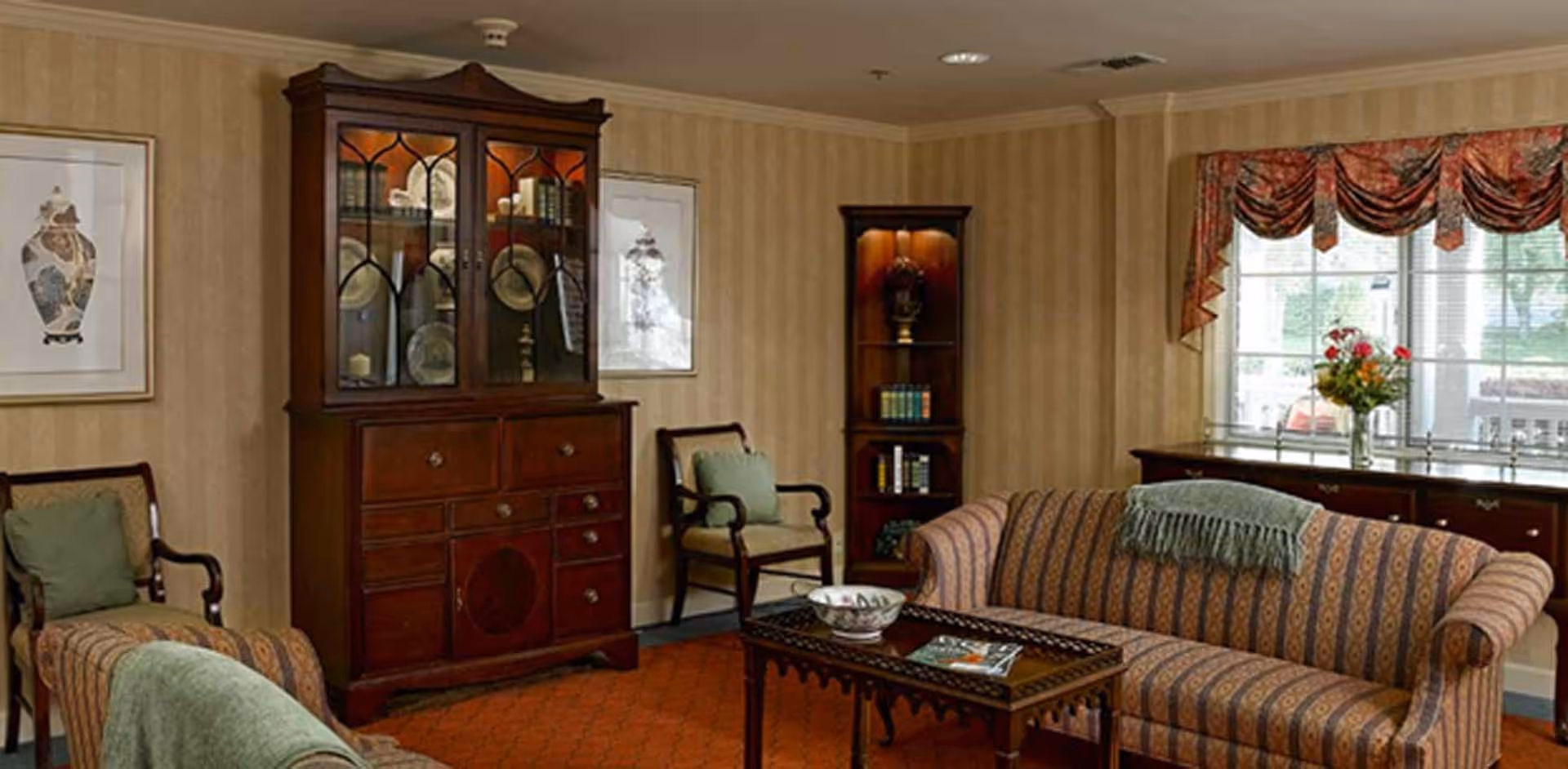 Comfortably furnished living room with patterned sofas, a wooden display cabinet, coffee table, and a window with a valanced curtain.