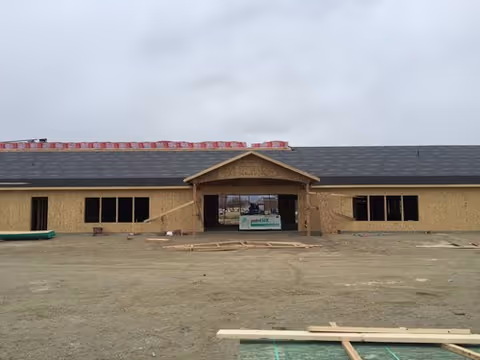 A single-story building under construction with wooden framing and sheathing. The roof is partially covered with black shingles and red roofing materials are stacked on top. The building has a central entrance with a small gabled roof and multiple window openings on either side. The ground in front is bare dirt with construction materials scattered around.