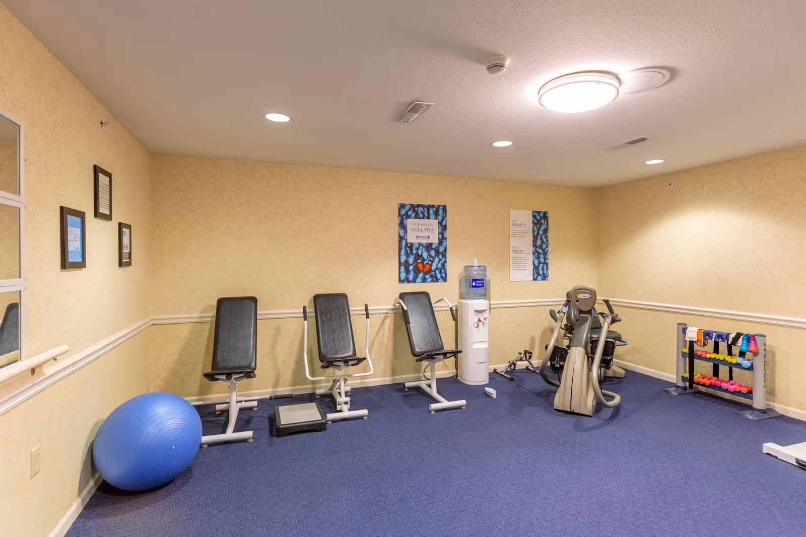 A small fitness room with exercise equipment including three seated workout machines, a stationary bike, a rack of colorful dumbbells, and a large blue exercise ball. The room has beige walls with framed motivational posters and a water cooler. The floor is covered with blue carpet.
