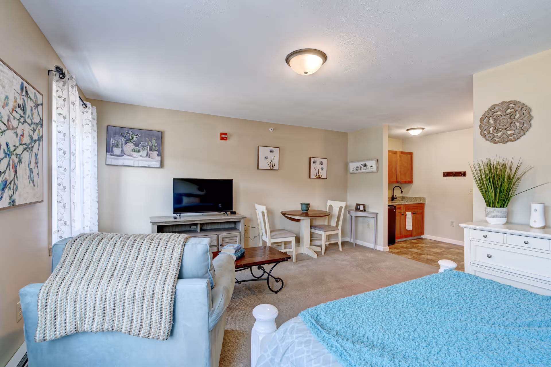 Studio-style living space with a bed and blue blanket in the foreground, a sofa, TV, small dining table, and a kitchenette in the background.