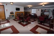 A common area with multiple tables and chairs arranged for seating. The room has a patterned floor with red and white tiles, bookshelves filled with books, a wall-mounted television, and ceiling lights. There are windows allowing natural light into the space.