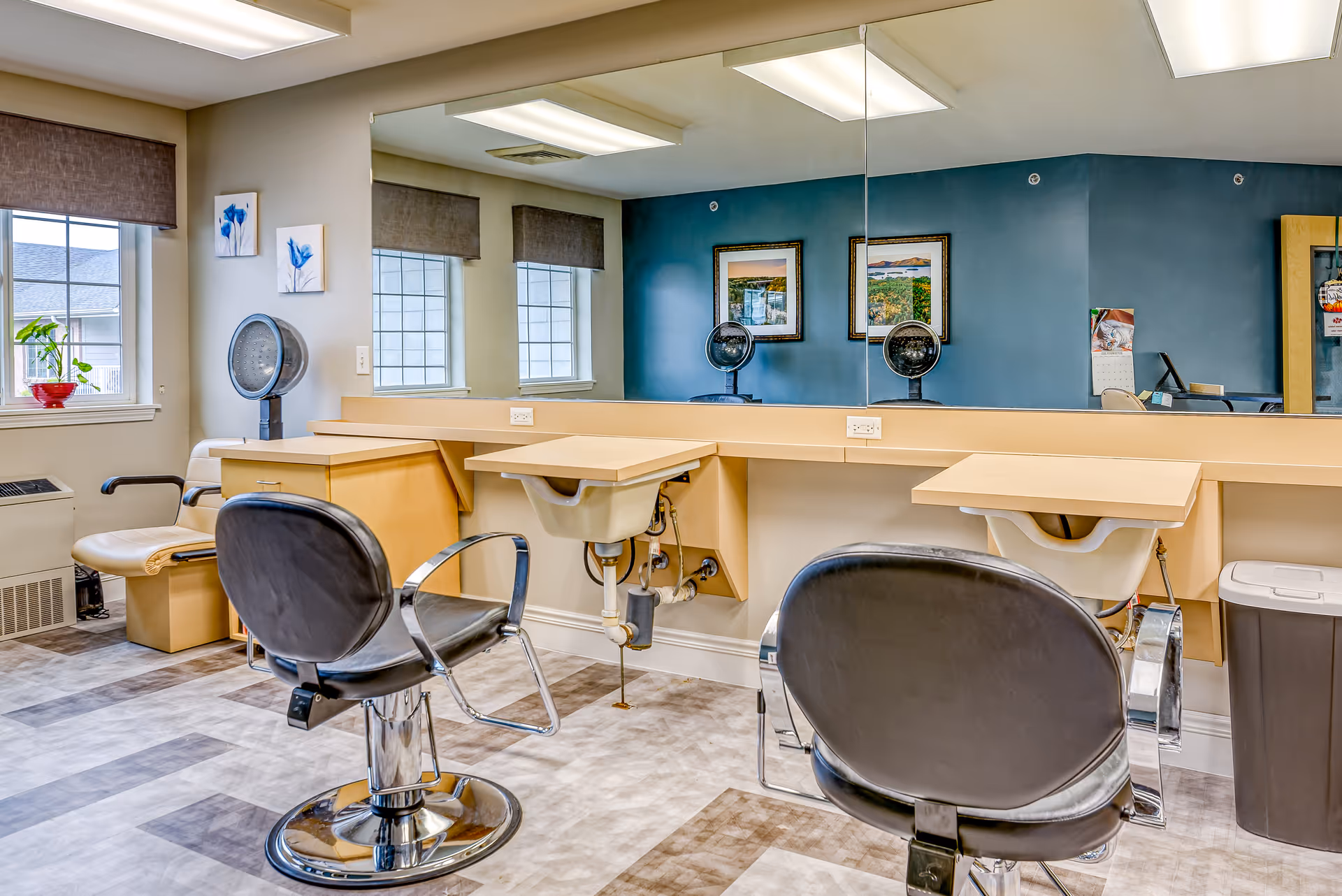 Interior of a salon area in a senior living facility with two black salon chairs facing a large mirror. There are two hair dryer stations with beige countertops and sinks underneath. The walls are painted blue and beige, with framed pictures hanging on the blue wall. A window with brown blinds and a small plant in a red pot is visible on the left side.