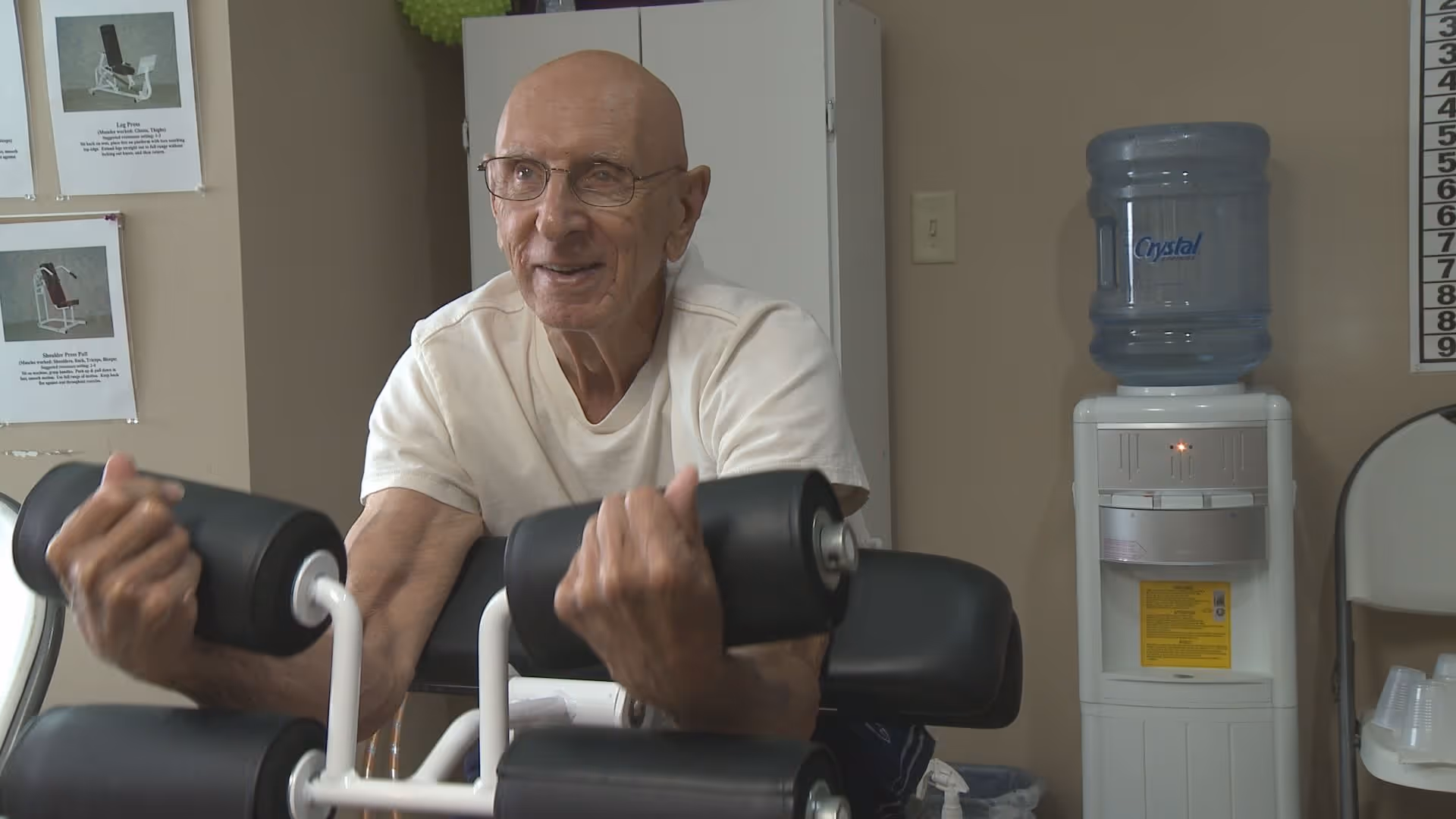An elderly man wearing glasses and a white t-shirt is exercising with a bicep curl machine in a room. Behind him, there is a water cooler, a white cabinet, and some exercise instruction posters on the wall.