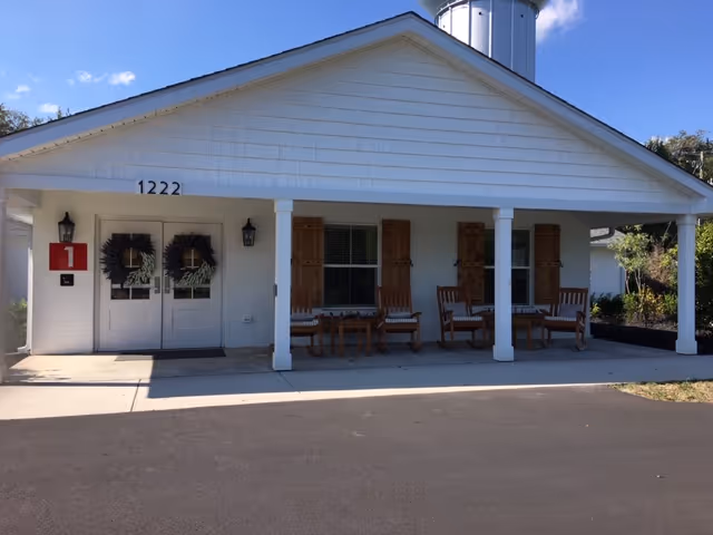 Front exterior view of a single-story building with white siding and a gabled roof. The entrance has double doors decorated with wreaths, flanked by two wall-mounted lanterns. There are wooden shutters on the windows and several wooden chairs and tables arranged on the covered porch. The building number 1222 is displayed above the doors. The sky is clear and blue.