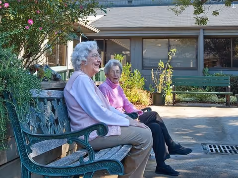 Two older women seated on a decorative bench chatting in a sunny courtyard outside a single-story assisted living building.