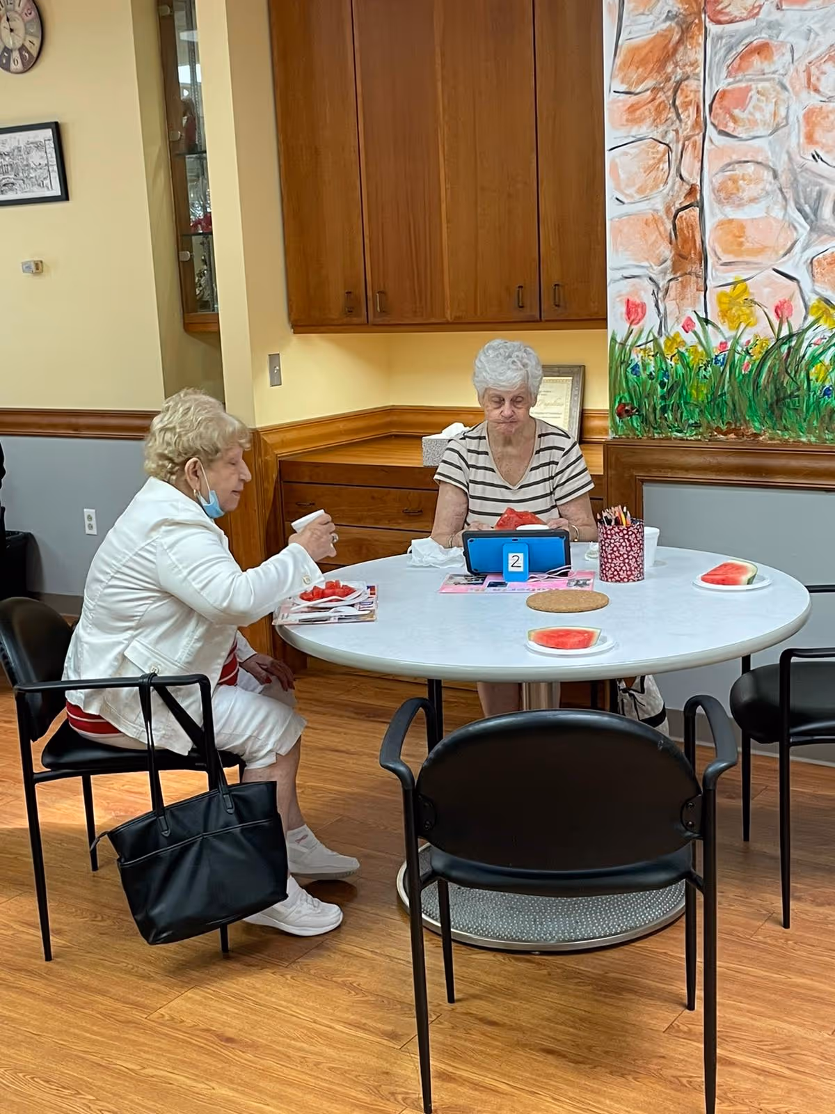 Two elderly women sitting at a round table in a room with wooden flooring and cabinets. One woman is eating watermelon while the other is holding a cup. The table has plates with watermelon slices, a container with colored pencils, and a small blue device with the number 2 on it. There is a colorful mural of flowers and stones on the wall behind them.