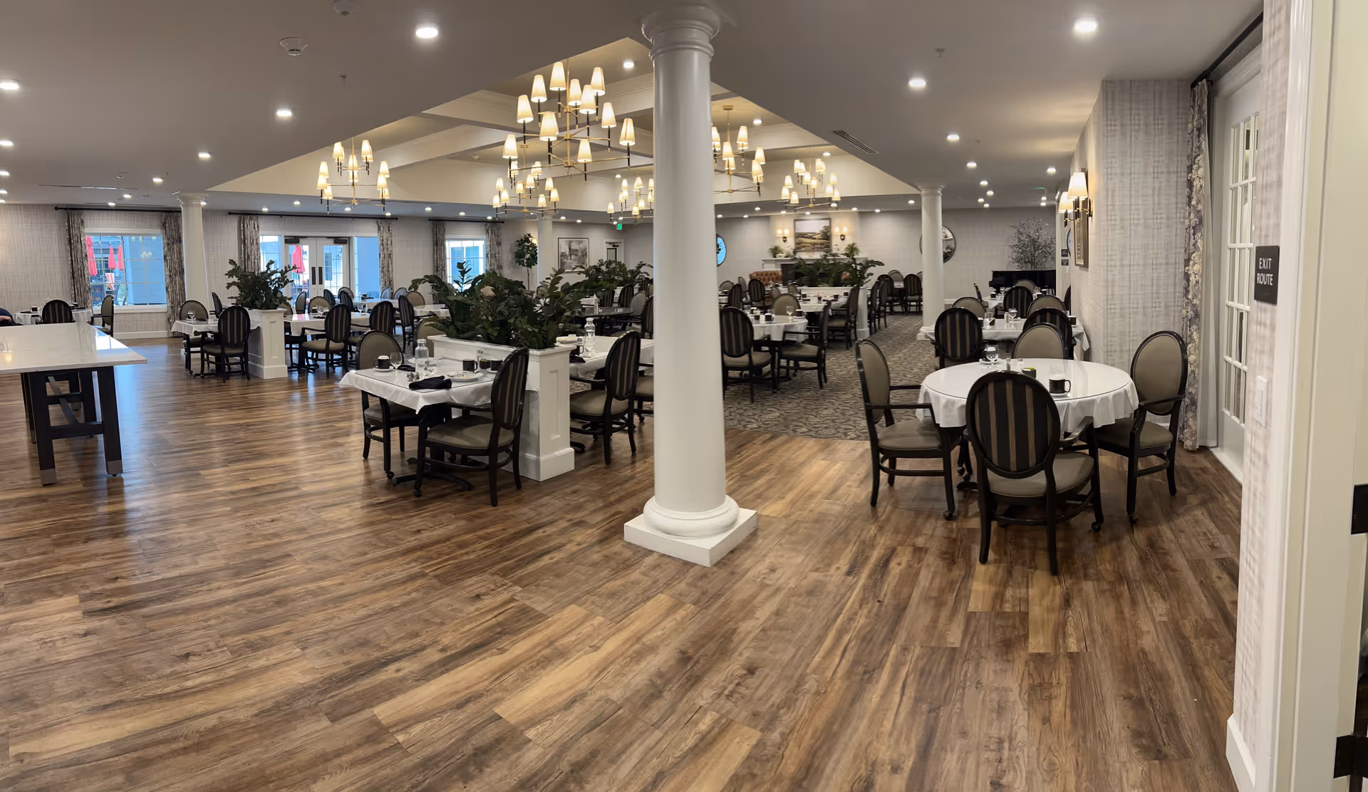 Spacious dining room in an assisted living community with multiple round and rectangular tables covered with white tablecloths, set with glassware and napkins. The room features wooden flooring, carpeted areas, white columns, large windows with curtains, and modern chandeliers hanging from the ceiling. There are plants placed on dividers between tables and an exit door on the right side.