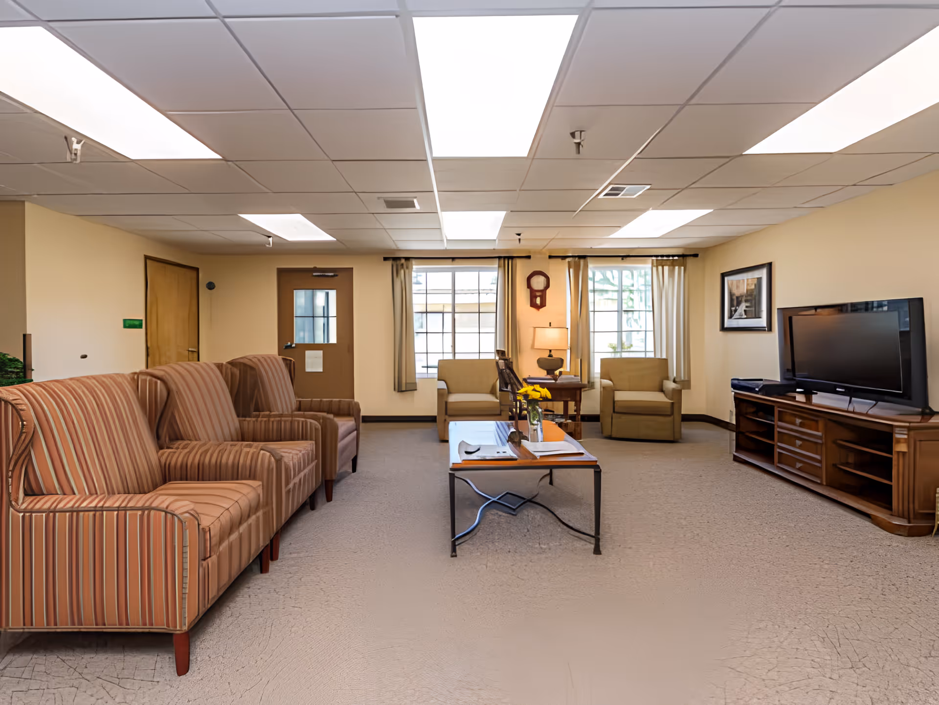 A well-lit living room with beige walls and carpeted floor. There are three striped armchairs on the left, two beige armchairs near the windows at the back, and a wooden coffee table with a vase of yellow flowers in the center. A wooden TV stand with a flat-screen television is on the right wall, and a framed picture hangs above it. The room has a door and two large windows with curtains, and a wall clock is mounted between the windows.