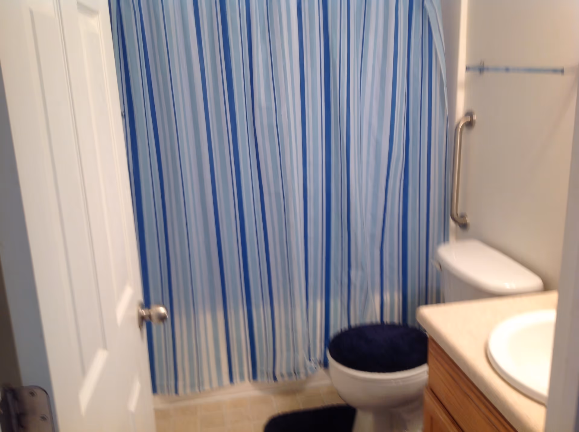 A small bathroom with a white door partially open, a toilet with a dark blue cover, a matching dark blue bath mat, a sink with a beige countertop, and a shower with a blue and white striped curtain. There is a metal grab bar on the wall next to the toilet.
