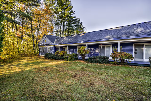Exterior view of a single-story blue building with white trim surrounded by green grass and trees with autumn foliage under a clear sky.
