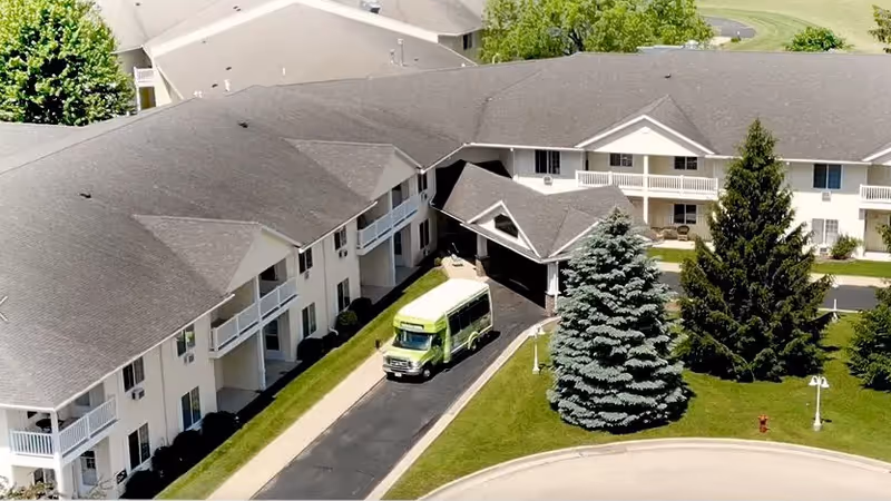 Aerial view of Charleston House Assisted Living facility showing a two-story building with balconies, a driveway with a green shuttle bus, and landscaped grounds with large evergreen trees.