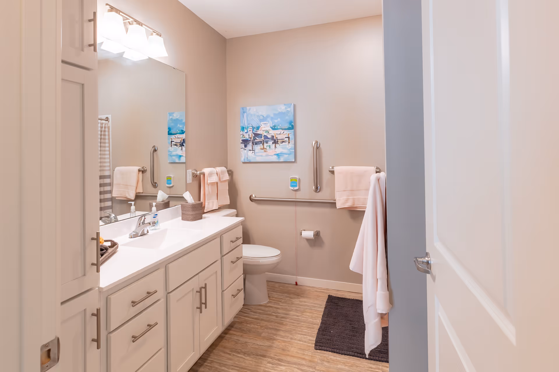 Accessible bathroom with a white vanity and sink, large mirror, toilet with grab bars, and pink towels.