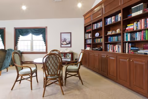 A cozy common room featuring a round table with four chairs, large built-in wooden bookshelves filled with books, and windows with green valances.