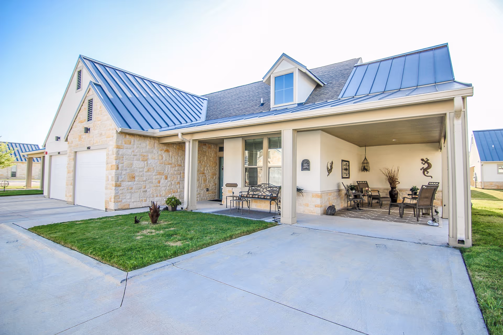 Exterior view of a residential building with a stone facade and blue metal roof. The building features a covered patio area with outdoor seating, decorative wall art, and a small grassy area in front. The driveway and garage doors are visible.