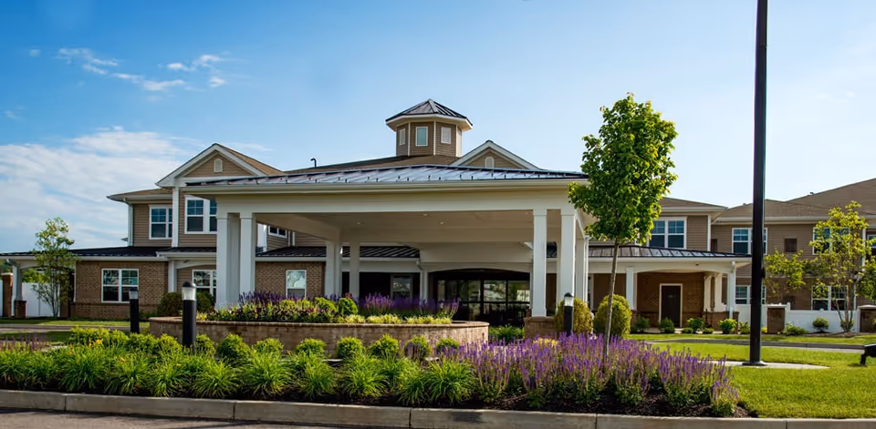 Front exterior view of Bellaire at Devonshire Assisted Living & Memory Care building with a covered entrance, landscaped garden with green shrubs and purple flowers, and a clear blue sky.